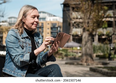 Mature Blonde Woman Using Her Smartphone Sitting On A Bench. She Is Wearing Denim Outfit.