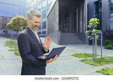 Mature Boss Talking Standing On Video Call, Senior Gray-haired Businessman Holding Netbook Looking At Web Camera And Explaining Discussing, Man In Business Suit Outside Office Building With Laptop