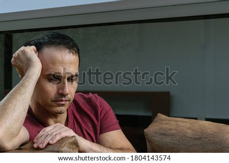 Mature Brazilian man (44 years old) sitting on the brown sofa, behind the glass, sad and crestfallen.