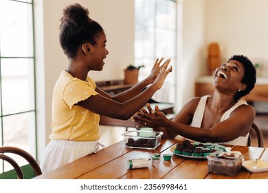 Mature Brazilian mom and her young daughter happily making brigadeiro together, laughing while rolling the chocolate balls into candy cups. Fun family moment between mother and child at home.