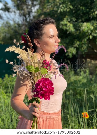A mature brunette woman with braided brown hair in a pink and cream T-shirt and a long skirt stands with a flower bouquet in the garden. Side view (profile) close-up