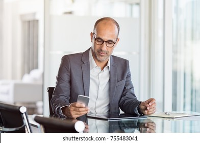 Mature Business Man In Formal Clothing Wearing Spectacles Using Mobile Phone. Serious Businessman Using Smartphone And Digital Tablet At Work. Manager In Suit Using Cellphone In A Modern Office.