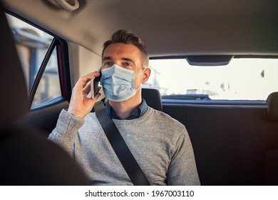 Mature Business Man Wearing A Face Mask For Safety Against Covid-19 Sitting In Car Rear Seat And Talking Over Phone. Man In A Business Call In Taxi Ride Wearing Face Mask For Precaution Against Corona