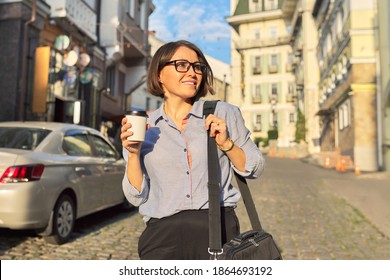 Mature Business Woman In Glasses With Office Laptop Bag Walking Along City Street. Happy Smiling Middle Aged Female With Cup Of Coffee, Urban Style Background