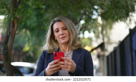 Mature Business Woman Holding Phone While Walking In City Street In Daylight. Female Person In 50s Reading Message On Smartphone Device In Urban Sidewalk