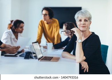 Mature Business Woman In Meeting Room With Colleagues Discussing In Background. Confident Female Professional At Board Room Meeting.