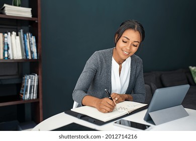 Mature business woman takes note of the appointments on the agenda while using digital tablet at home. Freelancer writing details in notebook while working in living room. African woman in smart work.