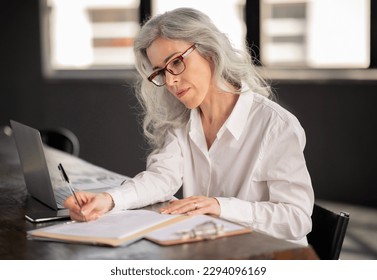 Mature Business Woman Taking Notes Near Laptop Computer Sitting At Desk In Modern Office, Wearing Glasses. Female HR Writing Doing Paperwork At Workplace. Entrepreneurship Career Concept - Shutterstock ID 2294096169