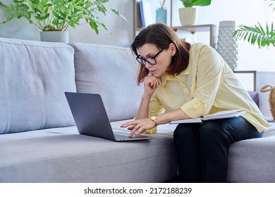 Mature business woman working on couch, using laptop making notes in notebook