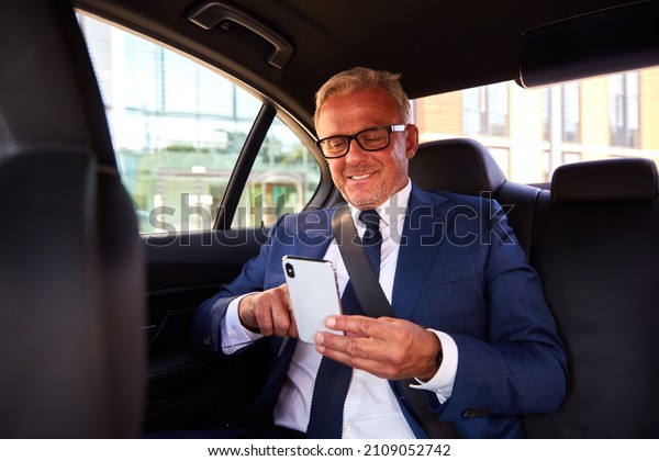 Mature Businessman In Back Of Taxi Or Car Checking\
Messages On Mobile Phone