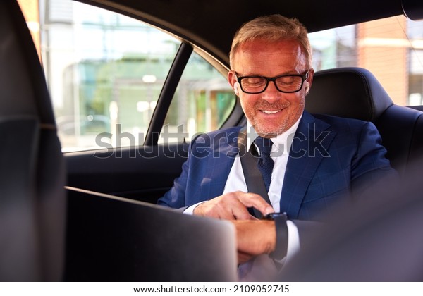 Mature Businessman In Back Of Taxi Or Car Working\
on Laptop Checking Smart\
Watch