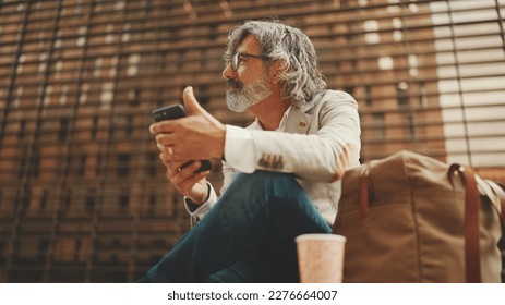 Mature businessman with beard in eyeglasses wearing gray jacket is using cell phone. Middle aged manager scrolling information on his smartphone while sitting outside the office - Shutterstock ID 2276664007