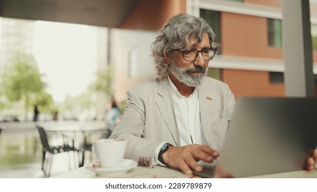 Mature businessman with beard in eyeglasses wearing gray jacket sits drinks coffee in cafe. Middle aged manager successful man working on laptop pc computer sit at cafe outdoors - Powered by Shutterstock