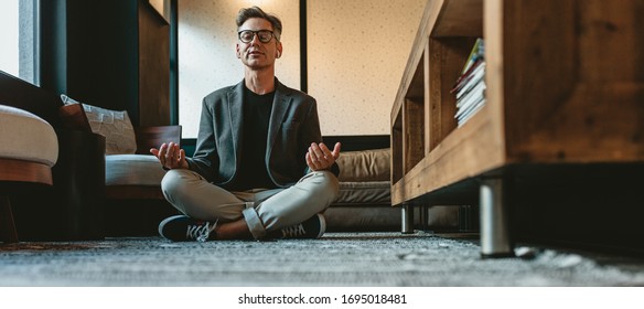 Mature businessman doing yoga meditation in office lounge. Male business executive meditating in lotus pose on the floor in the office.