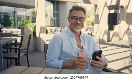 Mature businessman drinking cocktail, checking smartphone during break on sunny terrace. Portrait of happy man on summer vacation, smiling, relaxing. Leisure and effortless lifestyle. - Powered by Shutterstock