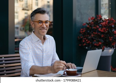 Mature Businessman Drinking Coffee In Cafe. Portrait Of Happy Handsome Man Wearing Stylish Eyeglasses Using Laptop, Looking At Camera, Smiling. Coffee Break Concept	