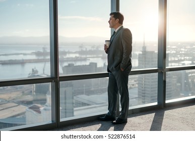 Mature businessman drinking a coffee and looking out of a window at the city skyline from an office building - Powered by Shutterstock