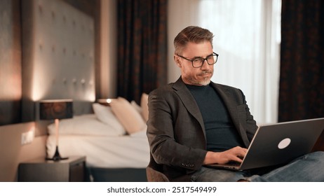 Mature businessman focuses on work in modern hotel room interior. Serious male professional working at home with computer. Mid adult man thinking. Entrepreneur managing business online. - Powered by Shutterstock