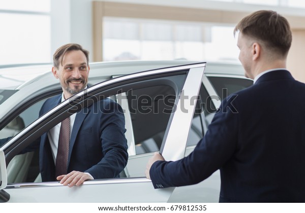 Mature businessman getting into his newly bought\
car with the salesman holding the door for him service\
professionalism luxury business seller retail rental transportation\
buying success concept