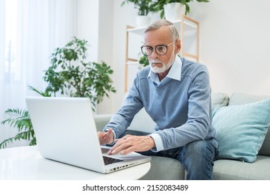 Mature Businessman In Glasses Works Online On Laptop Sitting On Sofa At Home. Focused Elderly Man In Elegant Outfit Deals With New Project Of Company