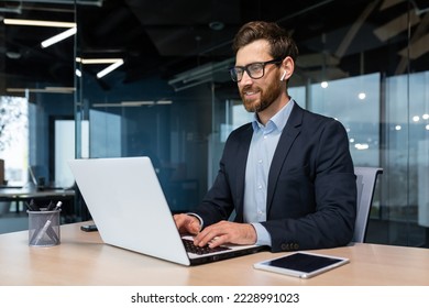 Mature businessman in headphones small earbuds talking on a video call using a laptop, boss at work at the desk in a business suit in the middle of the office smiling friendly. - Powered by Shutterstock