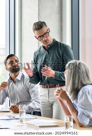 Mature businessman leader mentor talking to diverse colleagues team listening to caucasian ceo. Multicultural professionals project managers group negotiating in boardroom at meeting. Vertical.