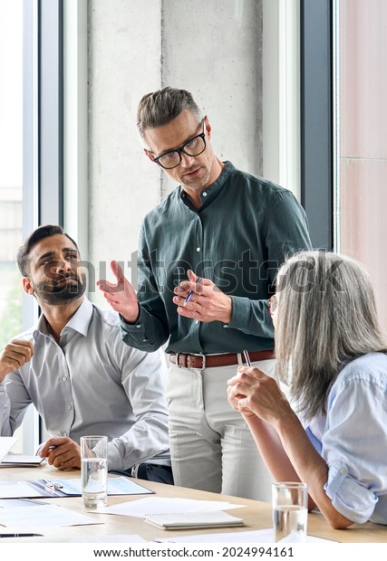 Mature businessman leader mentor talking to\
diverse colleagues team listening to caucasian ceo. Multicultural\
professionals project managers group negotiating in boardroom at\
meeting. Vertical.