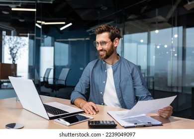 Mature businessman in shirt doing paperwork, man working with documents, contracts and bills sitting at table using laptop at work, financier accountant with beard and glasses.