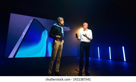 Mature Businessman Showing New Cellphone And Speaking With Bearded Engineer While Standing Against LED Screen During Presentation On Stage