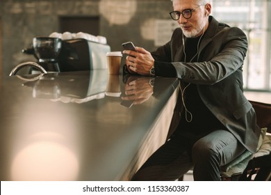 Mature businessman sitting at a cafe using mobile phone. Man in suit wearing earphones looking at his cellphone while relaxing in coffee shop. - Powered by Shutterstock