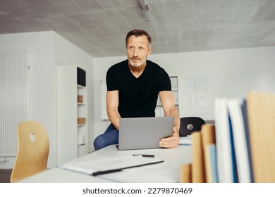 Mature Businessman Sitting on Desk in Office with Laptop and Looking Thoughtful at Something Off-Camera - Powered by Shutterstock