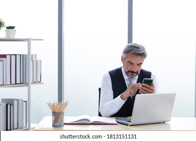 Mature businessman with smartphone in the office. Business people and technology concept. Serious businessman looking at smartphone in office. Portrait of executive on office desk using smartphone.