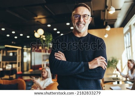 Mature businessman standing in a co-working space. Happy mature businessman smiling at the camera while standing with his arms crossed. Experienced entrepreneur working in a modern workspace.