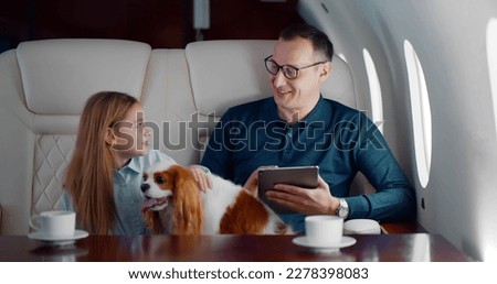 Mature businessman travelling on personal jet with little daughter and dog. Father using digital tablet and talking to preteen girl sitting in first class airplane interior
