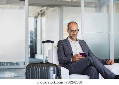 Mature businessman using mobile phone at the airport in the waiting room. Business man typing on smartphone in lounge area. Portrait of latin man sitting and holding passport with luggage. - Powered by Shutterstock