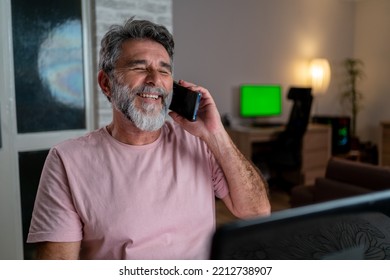 A Mature Businessman Working At Home With A Laptop On The Desk. Smiling Handsome Freelancer Working Remotely From Home. He Is Speaking On The Phone. Middle-aged Guy Working On Laptop