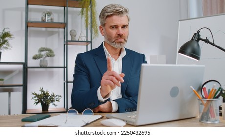 Mature businessman working on laptop computer shakes finger and saying No be careful scolding and giving advice to avoid danger mistake disapproval sign at office. Confident freelancer middle aged man