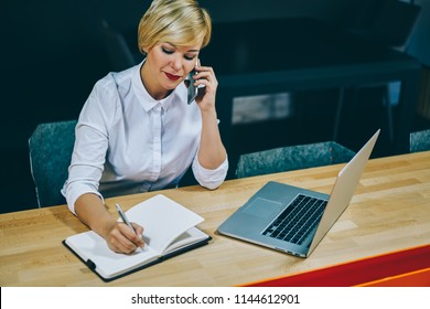 Mature Businesswoman Calling On Smartphone And Making Notes In Notepad During Working Process At Netbook With 4G Internet.Female Professor 50 Years Old Writing Information During Phone Conversation