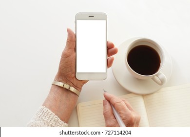 Mature Businesswoman Handwriting In Copybook Using Mobile With Blank White Copy Space Display For Your Information. Close Up Of Senior Woman's Hands Holding Cell Phone; Mug And Notebook On Desk