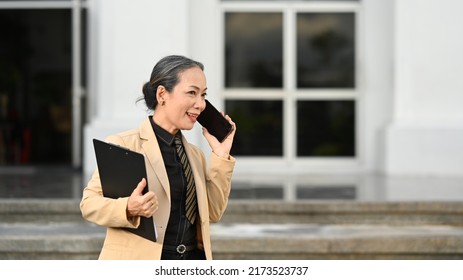 Mature Businesswoman Having Phone Conversation And Standing Outside Modern Office Building