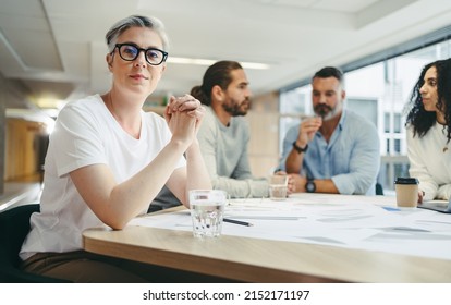 Mature businesswoman looking at the camera while sitting in a meeting with her team. Group of creative design professionals discussing a set of blueprints while working on a new project.