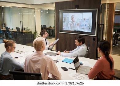 Mature Businesswoman O The Screen Holding The Online Conference For Business People Who Sitting At The Table And Listening To Her At Office