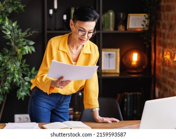 Mature businesswoman in smart casual clothes and glasses with documents leaning on table and reading data on laptop during work in home office with brick walls