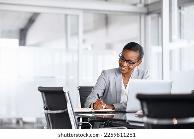 Mature businesswoman taking notes in notebook while using laptop at office. Mid adult black woman entrepreneur writing details on paper while working on laptop. Smiling african american business woman