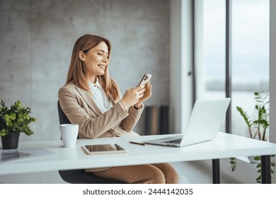 Mature businesswoman talking with somebody while sitting at office desk and working on laptop. Happy business woman chatting on digital gadgets online, holding mobile phone, using laptop 