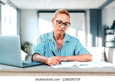 Mature Bussines Woman Working At Office. Writting In A Notebook While Sitting In Front Of A Laptop. Mature Accountant With Serious Face Expression Writting Down Checklist.
