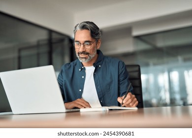 Mature busy businessman writing notes watching online webinar sitting at desk. Middle aged older professional business man using laptop working looking at computer technology in office. - Powered by Shutterstock