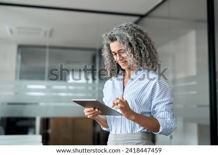 Mature busy happy businesswoman bank manager, older female corporate executive holding digital tablet standing at work. Middle aged professional business woman using tab computer in office.