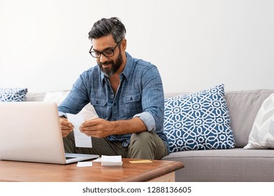 Mature Casual Man Using Laptop While Looking At Invoice. Smiling Latin Man Managing Finance With Bills And Laptop While Sitting On Couch At Home. Hispanic Guy Reading Expenses Wearing Eyeglasses.