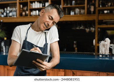 Mature caucasian barista waiter cafe restaurant owner talking on cellphone with customers, taking orders for takeout takeaway food delivery writing in clipboard near bar counter - Powered by Shutterstock